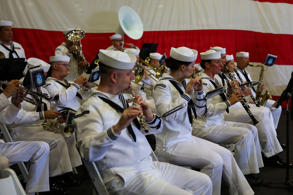 A U.S. Navy military band is performing in full dress white uniforms in front of a large American flag backdrop. The ensemble includes multiple musicians playing a variety of brass and woodwind instruments while seated in rows. The setting appears to be an indoor or tented ceremonial event.