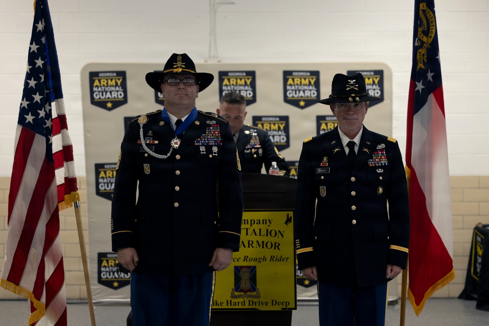 Two U.S. Army soldiers in dress uniforms stand at attention during a formal military ceremony for the Georgia Army National Guard. The soldier on the left is wearing a large medal (appears to be the Meritorious Service Medal or similar decoration) and a campaign/cavalry hat, suggesting this is a retirement or awards ceremony. A yellow unit sign referencing an Armor Battalion is visible between them, flanked by the U.S. flag and the Georgia state flag.