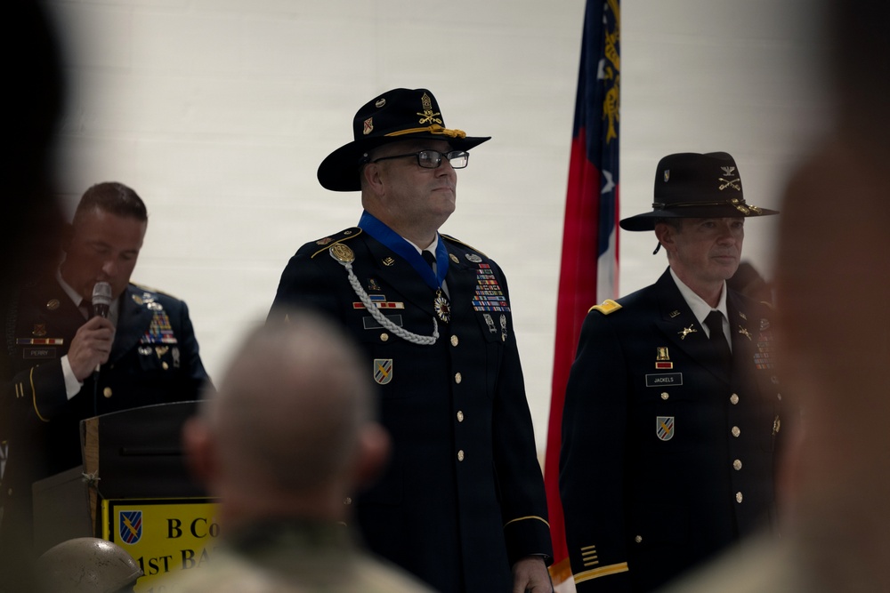 A formal U.S. Army military ceremony is taking place, likely a change of command or retirement ceremony for B Company, 1st Battalion. The central figure is a senior non-commissioned officer or officer wearing dress blues with a distinctive cavalry Stetson hat, a blue Medal of Honor or distinguished service medal around his neck, and an aiguillette. A second officer in cavalry Stetson stands to the right, and a third soldier speaks at a podium to the left.