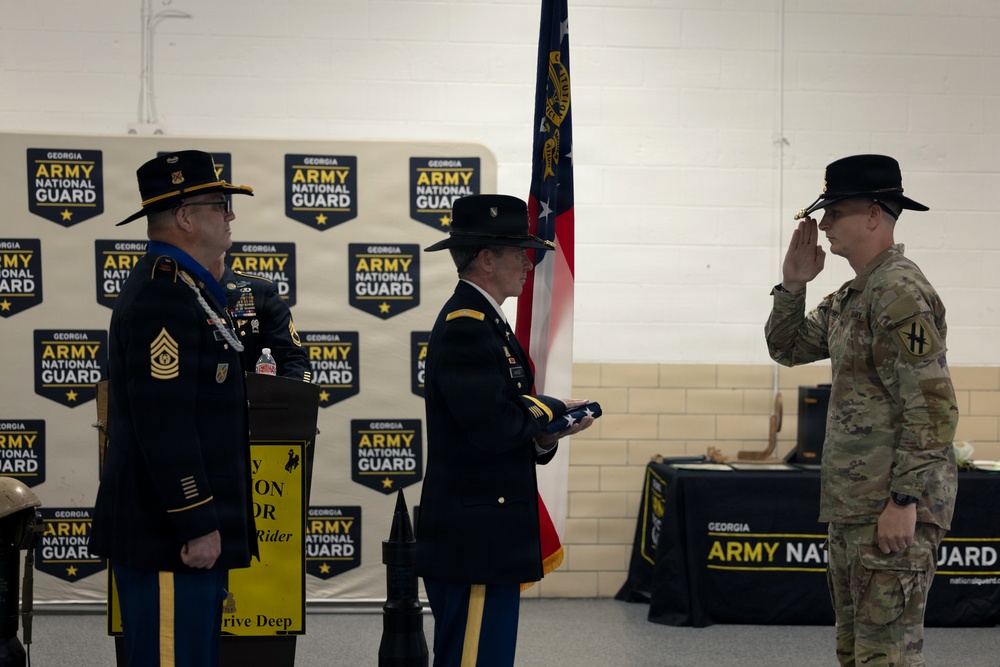 A formal Georgia Army National Guard military ceremony is taking place indoors, likely a retirement or awards ceremony. Three uniformed soldiers are visible, with one holding a folded American flag and another rendering a salute. The backdrop features repeated Georgia Army National Guard logos and a state flag is visible in the background.