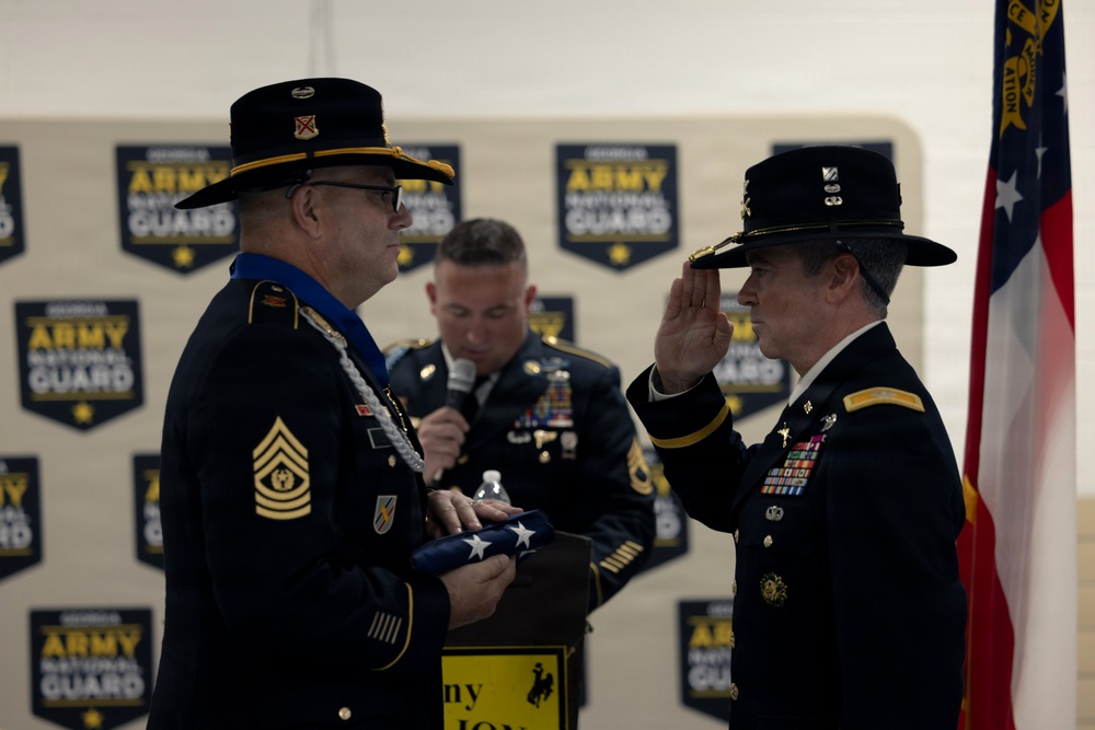 A formal U.S. Army National Guard military ceremony is taking place, likely a retirement or promotion event. Two senior soldiers in dress uniforms are at the forefront — one holding a folded flag and the other raising his right hand, suggesting an oath of office or formal presentation. A third soldier in the background appears to be reading from a script or speaking into a microphone.