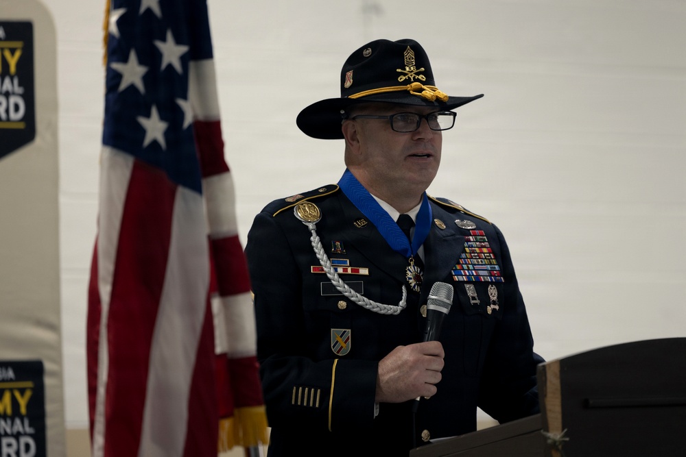 A senior U.S. Army officer in dress uniform is speaking at a podium during a formal military ceremony. He is wearing the Medal of Honor around his neck on a blue ribbon, along with numerous service ribbons, badges, and a cavalry-style campaign hat. An American flag and an Army National Guard banner are visible in the background.