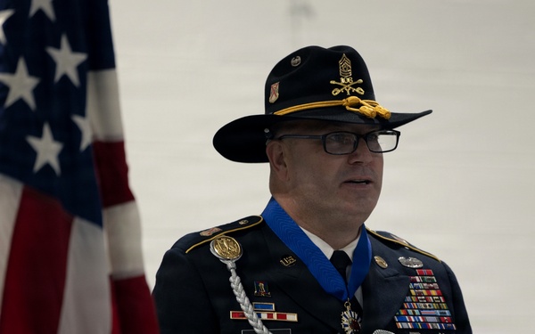 A senior U.S. Army officer in dress uniform is speaking at a podium during a formal military ceremony. He is wearing the Medal of Honor around his neck on a blue ribbon, along with numerous service ribbons, badges, and a cavalry-style campaign hat. An American flag and an Army National Guard banner are visible in the background.