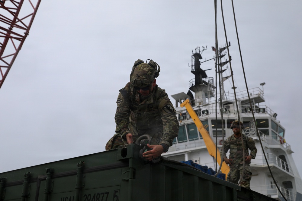 Military personnel in camouflage uniforms are conducting cargo loading or unloading operations at a port facility. A soldier in the foreground is securing or attaching rigging hardware to a green military container, while a second soldier stands nearby observing. A white vessel and a red crane are visible in the background.