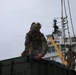 Military personnel in camouflage uniforms are conducting cargo loading or unloading operations at a port facility. A soldier in the foreground is securing or attaching rigging hardware to a green military container, while a second soldier stands nearby observing. A white vessel and a red crane are visible in the background.