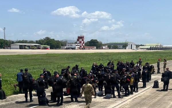 A large group of personnel dressed predominantly in black uniforms and tactical gear are assembled on an airport tarmac or taxiway. One individual in military camouflage uniform is walking toward the group. The setting appears to be a tropical or subtropical airfield, identifiable by the red-and-white checkered control tower and lush green vegetation in the background.