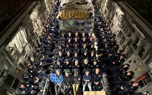 A large group of personnel, predominantly wearing matching dark navy uniforms with patches, are seated inside the cargo hold of a military transport aircraft, likely a C-17 Globemaster III. The photo is taken from an elevated angle looking down the length of the aircraft toward the tail ramp. An American flag is visible at the bottom of the frame near the open ramp area, and cargo is secured in the center of the aircraft.
