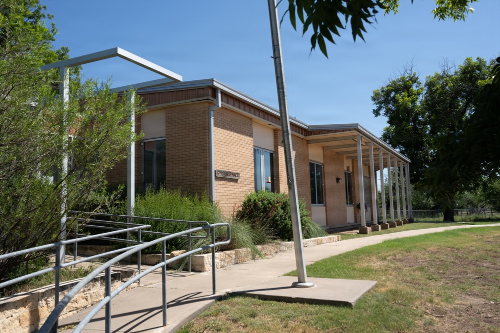 A single-story brick building with mid-century modern architectural features, including a covered walkway with metal posts along its facade. The building appears to be a public or institutional facility, identifiable by an 'ENTRANCE' sign and ADA-compliant accessibility ramp with metal railings. The surrounding landscape includes mature trees, shrubs, and a concrete sidewalk under a clear blue sky.