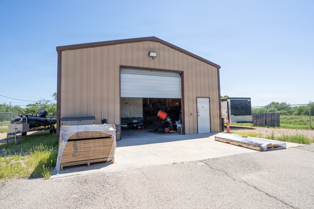 A metal pole barn or commercial workshop building with tan/beige corrugated steel siding and brown trim, photographed on a clear sunny day. The roll-up garage door is open, revealing equipment and tools inside. The property appears to be an active work or storage facility with materials staged outside.