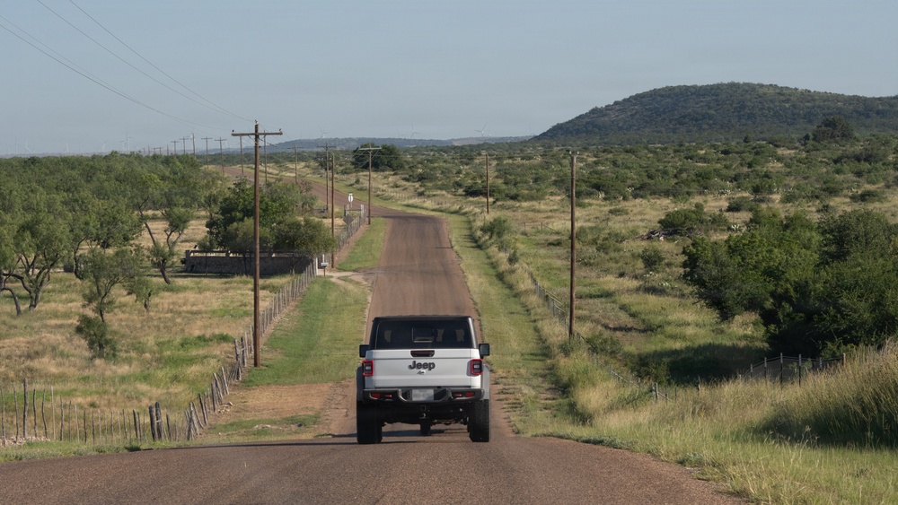 A white Jeep pickup truck (Gladiator) is driving away from the camera on a straight, narrow rural road through open scrubland terrain. The road stretches toward a distant mesa or hill covered in vegetation, flanked by wooden utility poles and wire fencing. Wind turbines are faintly visible on the horizon to the left, suggesting a rural area in the American Southwest, likely Texas.