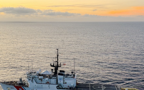 An aerial photograph of a U.S. Coast Guard cutter underway at sea, captured during sunset or sunrise conditions. The vessel bears the hull number 901 and the text 'U.S. COAST GUARD' prominently on its hull. The ship is actively moving through calm open ocean waters, leaving a visible wake.