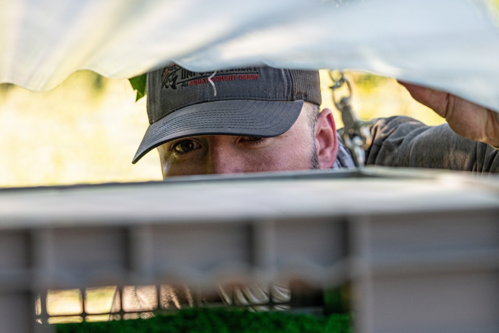 A man wearing a dark baseball cap is peering into or under a covered structure, likely a cage or crate with a white tarp or cover. The image is shot from a low angle inside the structure, looking up at the man's face framed between the cover and the edge of the container. The cap has embroidered text and a fish logo, suggesting a fishing or outdoor recreation theme.