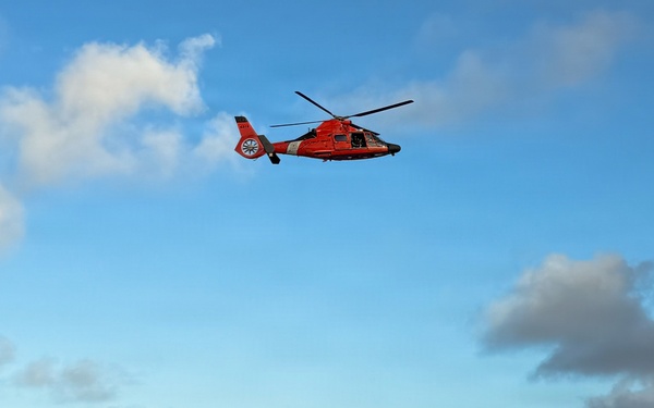 A U.S. Coast Guard helicopter hovers above a small white boat in open ocean waters. Several people are visible standing on the boat, suggesting a search and rescue or interdiction operation is underway. The scene appears to be taking place in calm but deep blue open sea conditions.