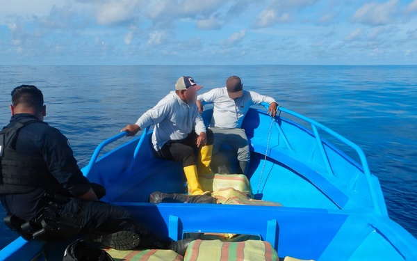 A blue open boat on open ocean contains multiple large bundled packages wrapped in striped woven sacks. One individual wearing a tactical vest and gloves, with partial text visible on their back, is present alongside two other individuals seated toward the bow. The scene appears consistent with a maritime law enforcement interdiction.