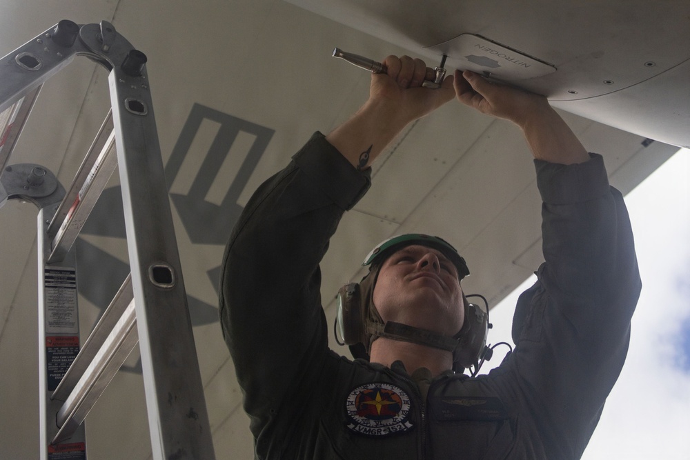 A military aviation technician or mechanic is performing maintenance work on the underside of an aircraft, using a hand tool to work on a panel labeled 'NITROGEN'. The individual is wearing a flight suit, green helmet, and hearing protection headset, and is using an aluminum ladder for access. The patch on their sleeve indicates unit designation 'VMGR-252', suggesting U.S. Marine Corps affiliation.