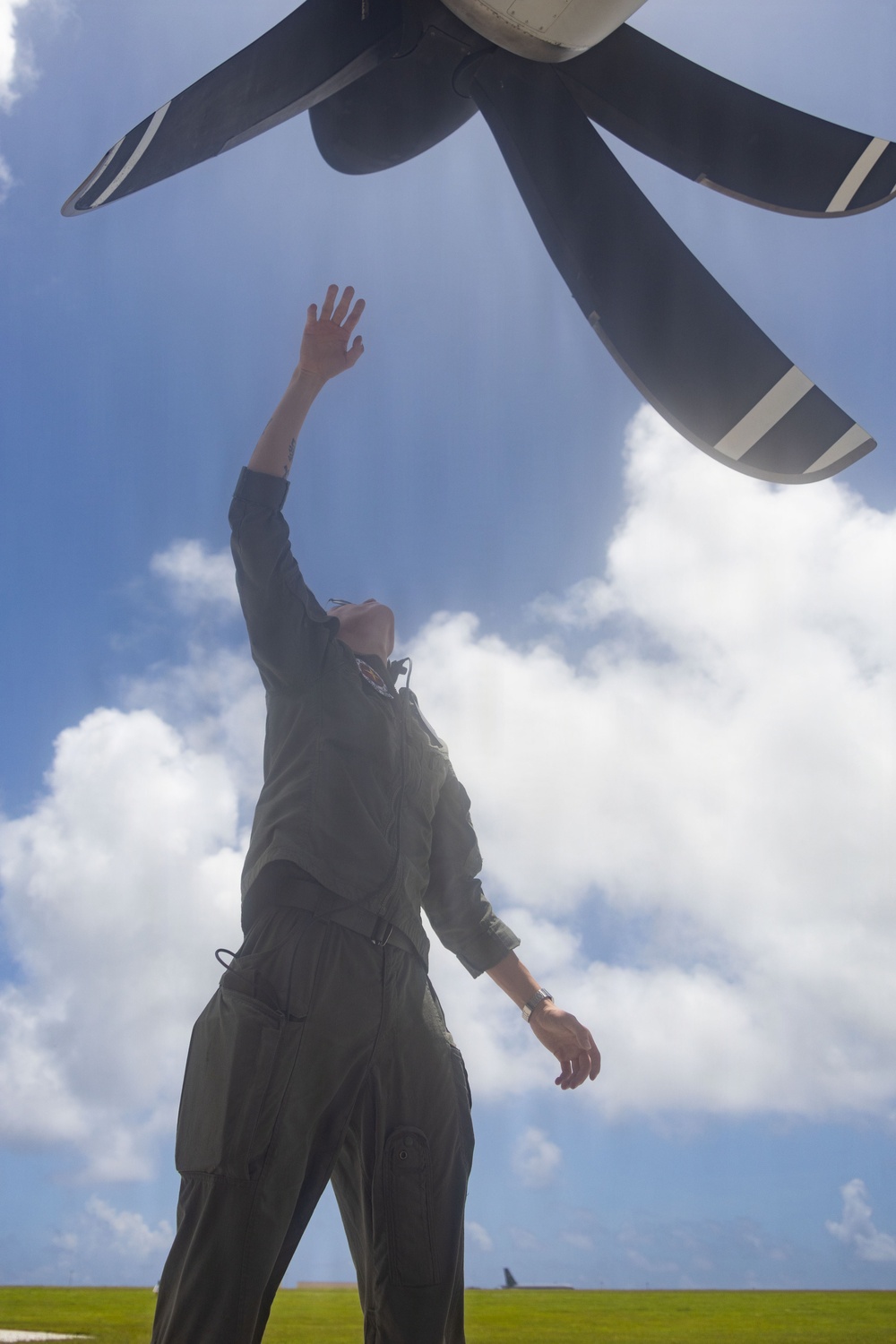 A person wearing an olive/sage green flight suit stands beneath a large multi-blade aircraft propeller, reaching upward toward it against a partly cloudy blue sky. The low-angle upward perspective creates a dramatic composition emphasizing the scale of the propeller relative to the individual. The individual appears to be conducting a pre-flight or post-flight inspection of the propeller blades.