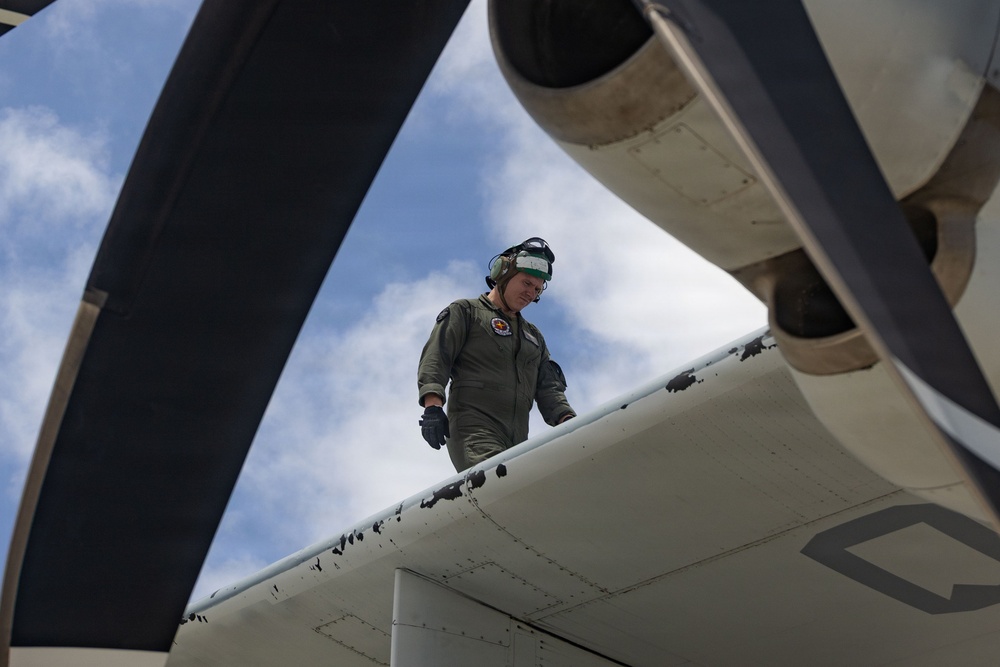 A military aviation crew member in an olive green flight suit is conducting a pre-flight or post-flight inspection on the wing of a turboprop aircraft. The photo is taken from a low angle beneath the aircraft's propeller, creating a dramatic perspective. The aircraft wing shows visible wear and dark scuff marks on its white painted surface.