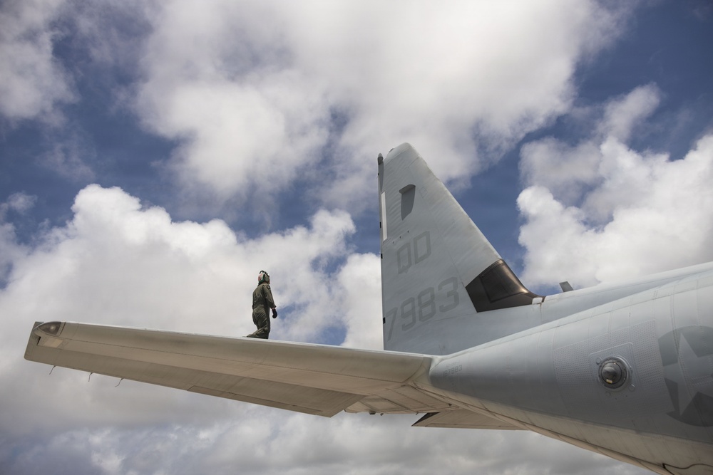 A military aircraft technician or crew member in a flight suit and helmet is standing on the horizontal stabilizer/tail wing of a large military aircraft. The tail section displays the markings 'DD' and '7883' on a light gray fuselage. The photo is taken from a low angle looking upward, with a partly cloudy blue sky as the backdrop.