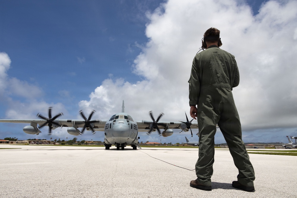 A military ground crew member in olive green flight suit and headset stands on a tarmac facing a C-130 Hercules turboprop aircraft with all four propellers spinning. The aircraft bears the tail number 983 and appears to be preparing for departure or undergoing engine run-up checks. The scene takes place at a military airfield under partly cloudy skies, likely in a tropical or subtropical location based on the vegetation visible in the background.