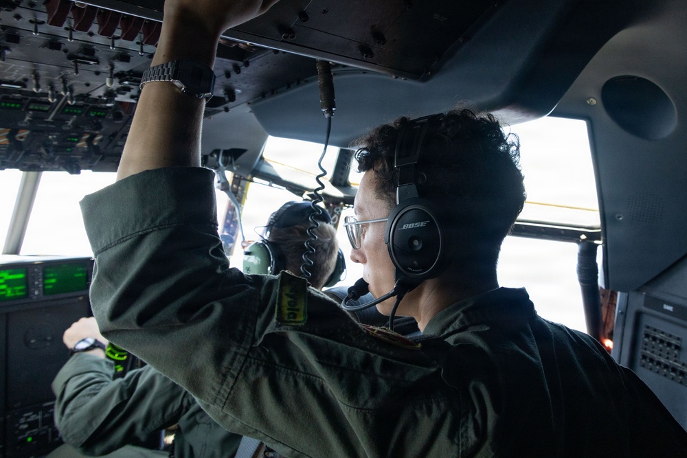 Two crew members in olive/flight suits are visible inside a military aircraft cockpit. The foreground crew member is wearing Bose aviation headsets and glasses while reaching up to manipulate overhead panel controls. Green avionics displays and complex overhead switch panels are visible, consistent with a large military transport aircraft.