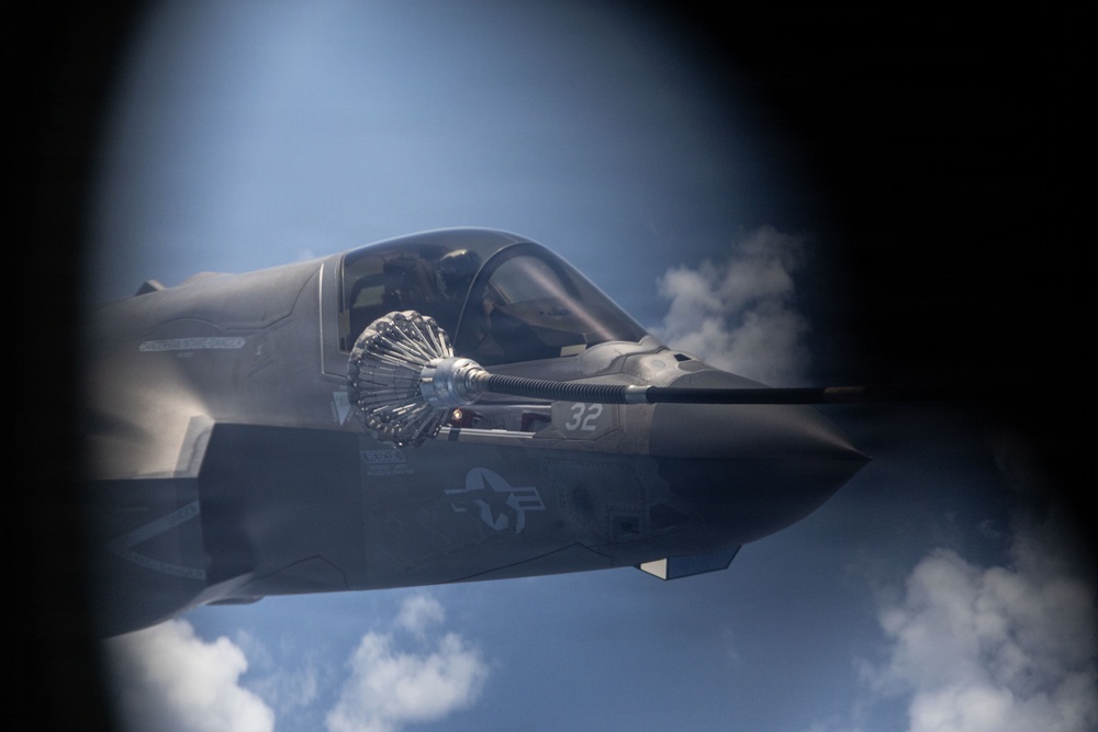 A U.S. military F-35 fighter jet, marked with the number '32' and a U.S. Air Force roundel, is captured during an aerial refueling operation. A refueling drogue (basket) is visible connected to the aircraft's refueling probe, with vapor or fuel mist visible around the connection point. The image is shot from the perspective of the tanker aircraft, framed through what appears to be an observation window.