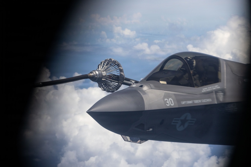 An aerial refueling operation is captured in flight, showing a military jet aircraft approaching a drogue basket attached to a refueling hose. The aircraft bears the number '30' and displays U.S. military insignia, consistent with a U.S. Marine Corps F-35B variant. The image is shot through what appears to be a porthole or observation window of the tanker aircraft, with clouds visible in the background.