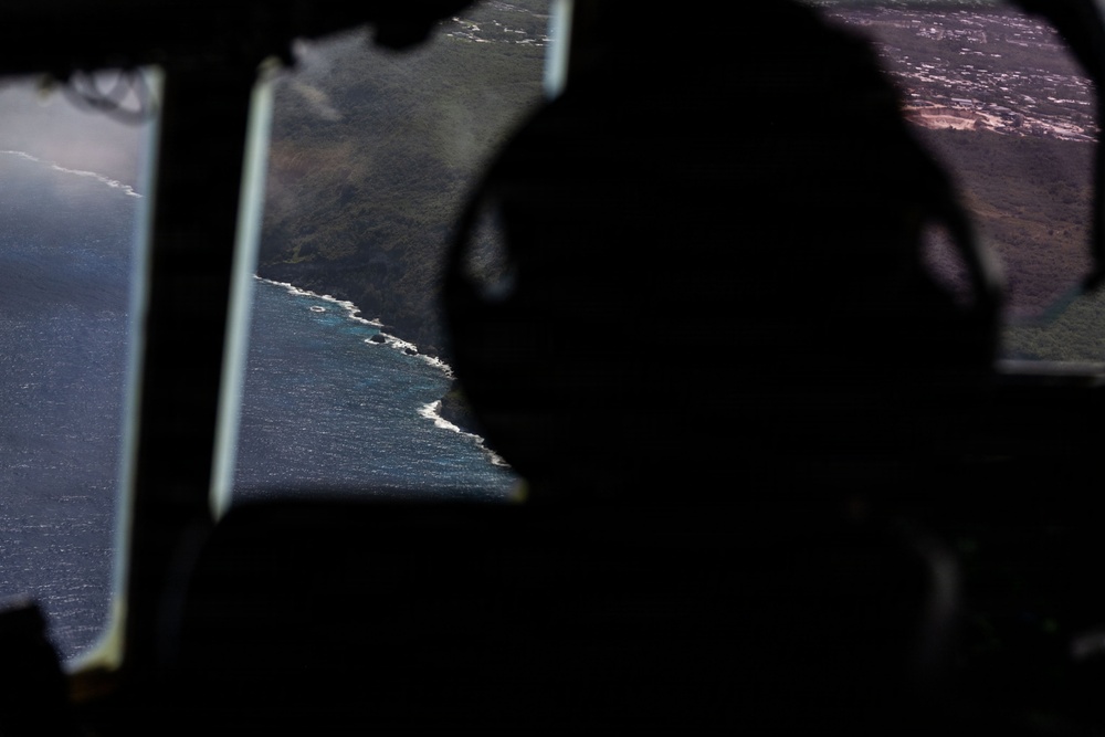 The image is taken from inside an aircraft cockpit or cabin, showing a silhouetted figure wearing what appears to be a headset looking out through windows at a coastal landscape below. The aerial view reveals a rugged coastline where dark volcanic or mountainous terrain meets turquoise ocean water with visible surf. The high contrast between the dark interior and bright exterior creates a dramatic silhouette composition.