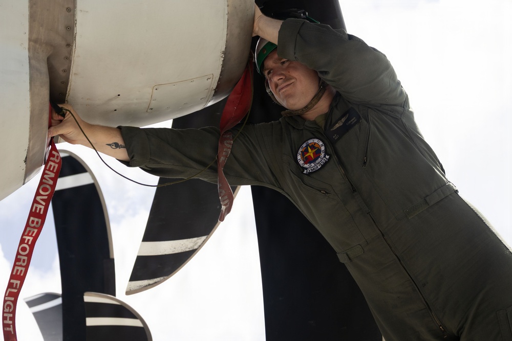 A military aviator or crew member in an olive green flight suit is removing a 'Remove Before Flight' safety streamer from an aircraft component, likely an engine intake or sensor cover. The individual is wearing a green helmet and displays a squadron patch on their flight suit. This appears to be a pre-flight inspection or preparation activity on a military aircraft.