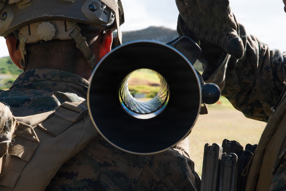 The image shows what appears to be U.S. Marines in digital camouflage (MARPAT) uniforms during a weapons training or maintenance exercise. The central focal point is a view down the muzzle of a large-caliber barrel, revealing the rifling (spiral grooves) inside the bore. The setting appears to be an outdoor training range with green hills visible in the background.