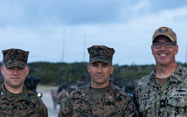 Three U.S. military personnel pose for a photo on what appears to be a tarmac or airfield. Two are wearing U.S. Marine Corps MARPAT woodland camouflage uniforms, while the third wears a U.S. Navy NWU Type III uniform. Military vehicles and equipment are visible in the background.