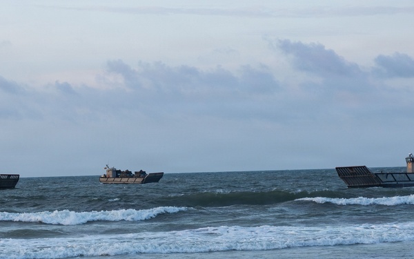 Three military landing craft are visible in coastal waters approaching or departing a sandy beach. The vessels appear to be utility landing craft (LCU) or mechanized landing craft (LCM) type, consistent with amphibious military operations. The scene is photographed from the shoreline under overcast, low-light conditions.