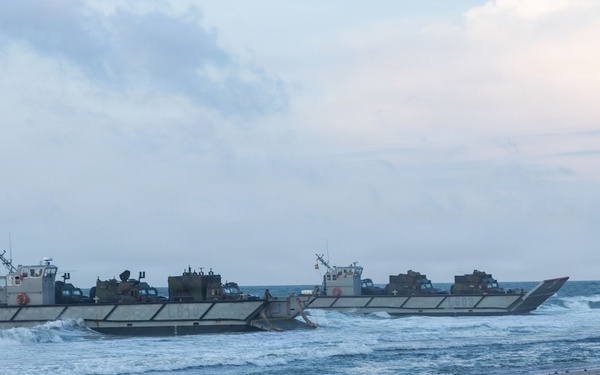 Two military landing craft are approaching or beaching on a sandy shoreline, each carrying multiple military vehicles on their open decks. The scene appears to depict an amphibious landing operation, likely a military exercise or training event. The overcast sky and choppy surf conditions are visible in the background.