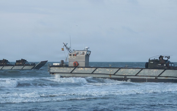 The image shows a military amphibious landing operation with multiple landing craft approaching a sandy beach through moderate surf. At least two landing craft utility (LCU) or similar flat-bottomed military vessels are visible, with their bow ramps lowered or in position for beaching. A military vehicle is visible on the rightmost craft, suggesting an active amphibious assault or exercise.