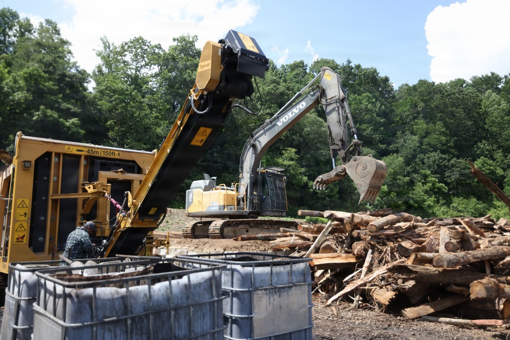An outdoor wood processing operation is underway, featuring a Volvo excavator with a grapple attachment handling logs from a large pile of cut timber. A yellow industrial machine, labeled with '45m/150ft' markings and a 'Tigercat' brand identifier, appears to be a wood chipper or grinder positioned to receive material. A worker in a plaid shirt is visible near the yellow machine, overseeing the operation.