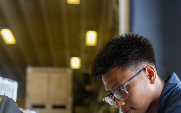 A young man wearing blue coveralls, clear-framed glasses, and black gloves is crouched down working on a large circular white metal component, likely a hatch or wheel mechanism. He has visible sleeve tattoos on his forearms and appears to be in an industrial or naval/military hangar environment. The image is a professional, sharp close-up photograph with strong depth of field.