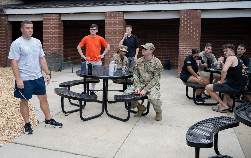 A group of approximately 10 individuals are gathered in an outdoor courtyard area adjacent to a brick building. The group includes both military personnel in camouflage uniforms and civilians in casual clothing, appearing to be engaged in a briefing or informal discussion. One person in a gray t-shirt with what appears to be a military logo stands apart, seemingly addressing the group.