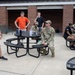 A group of approximately 10 individuals are gathered in an outdoor courtyard area adjacent to a brick building. The group includes both military personnel in camouflage uniforms and civilians in casual clothing, appearing to be engaged in a briefing or informal discussion. One person in a gray t-shirt with what appears to be a military logo stands apart, seemingly addressing the group.