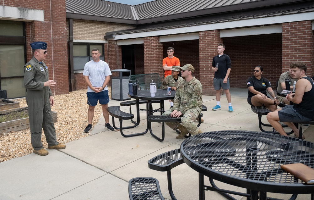 A military officer in a flight suit is addressing a small group of personnel and civilians in an outdoor courtyard area of a brick building. The group includes individuals in military uniforms (OCP/camouflage pattern) and others in civilian casual clothing, seated and standing around outdoor metal picnic tables. The setting appears to be a military installation, likely during an informal briefing or meet-and-greet session.