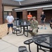 A military officer in a flight suit is addressing a small group of personnel and civilians in an outdoor courtyard area of a brick building. The group includes individuals in military uniforms (OCP/camouflage pattern) and others in civilian casual clothing, seated and standing around outdoor metal picnic tables. The setting appears to be a military installation, likely during an informal briefing or meet-and-greet session.