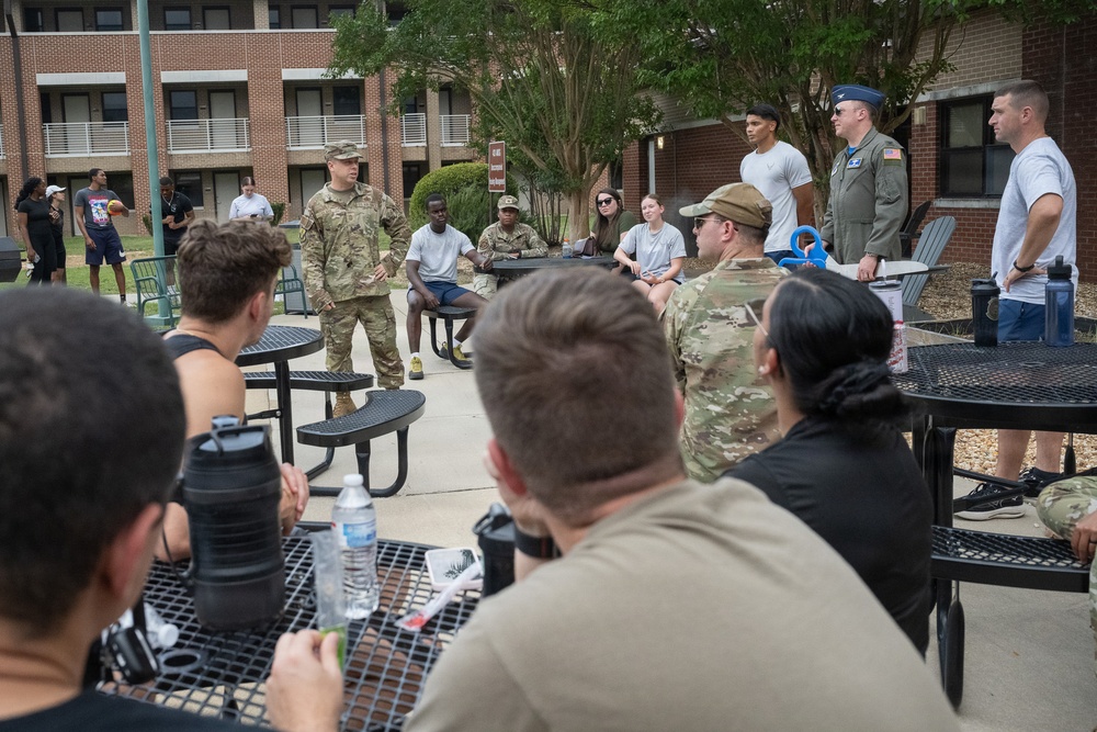 A group of military personnel and civilians are gathered outdoors in a courtyard area near a brick building, appearing to be in an informal briefing or group discussion. One individual in Army combat uniform (ACU/OCP pattern) appears to be addressing the assembled group. The setting suggests a military installation with a mix of uniformed and civilian attendees.