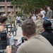 A group of military personnel and civilians are gathered outdoors in a courtyard area near a brick building, appearing to be in an informal briefing or group discussion. One individual in Army combat uniform (ACU/OCP pattern) appears to be addressing the assembled group. The setting suggests a military installation with a mix of uniformed and civilian attendees.