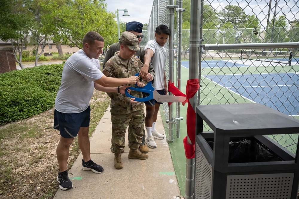 A ribbon-cutting ceremony is taking place at what appears to be a newly renovated or opened tennis court facility. Several individuals, including at least one person in military camouflage uniform and others in athletic wear, are using large ceremonial blue scissors to cut a red ribbon at the entrance gate. The setting appears to be on a military installation based on the uniforms and facility style.