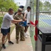 A ribbon-cutting ceremony is taking place at what appears to be a newly renovated or opened tennis court facility. Several individuals, including at least one person in military camouflage uniform and others in athletic wear, are using large ceremonial blue scissors to cut a red ribbon at the entrance gate. The setting appears to be on a military installation based on the uniforms and facility style.
