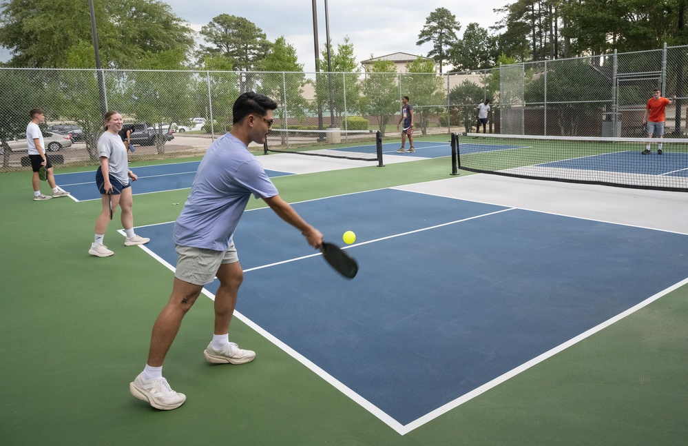 A group of young adults are playing pickleball on multiple outdoor courts with blue and green surfaces. The foreground shows a man in a light blue shirt mid-swing with a pickleball paddle, about to hit a yellow ball. Additional players are visible on adjacent courts in the background.