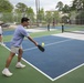 A group of young adults are playing pickleball on multiple outdoor courts with blue and green surfaces. The foreground shows a man in a light blue shirt mid-swing with a pickleball paddle, about to hit a yellow ball. Additional players are visible on adjacent courts in the background.