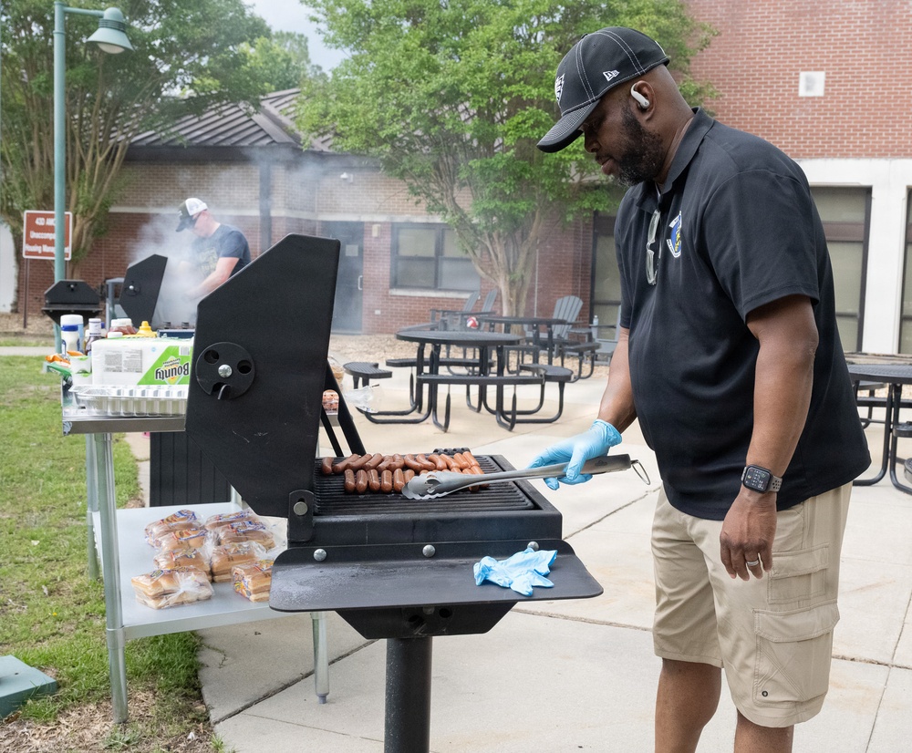 A man wearing a black polo shirt, black cap, and blue nitrile gloves is grilling hot dogs on an outdoor charcoal grill. The setting appears to be a community or institutional outdoor area with picnic tables, brick buildings, and trees in the background. A second person is visible in the background also tending to another grill, suggesting a group cookout or community event.