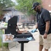 A man wearing a black polo shirt, black cap, and blue nitrile gloves is grilling hot dogs on an outdoor charcoal grill. The setting appears to be a community or institutional outdoor area with picnic tables, brick buildings, and trees in the background. A second person is visible in the background also tending to another grill, suggesting a group cookout or community event.