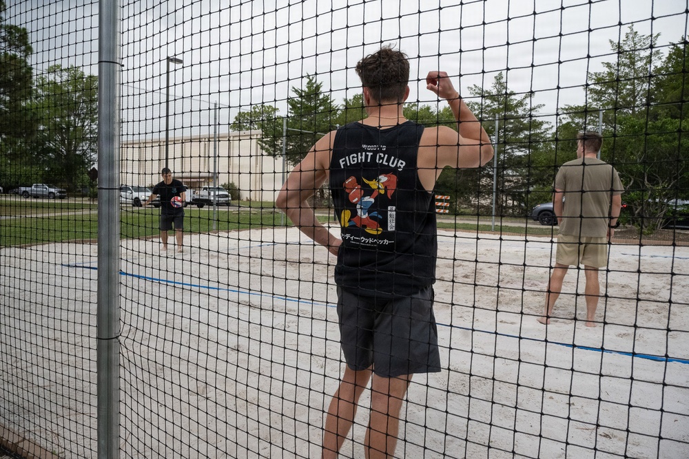 Three individuals are playing beach volleyball on a sand court, viewed through a black mesh net. The central figure wears a black sleeveless shirt reading 'Fight Club' with a cartoon graphic and Japanese text. The setting appears to be an outdoor recreational facility with trees, parked vehicles, and a building visible in the background.
