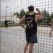 Three individuals are playing beach volleyball on a sand court, viewed through a black mesh net. The central figure wears a black sleeveless shirt reading 'Fight Club' with a cartoon graphic and Japanese text. The setting appears to be an outdoor recreational facility with trees, parked vehicles, and a building visible in the background.