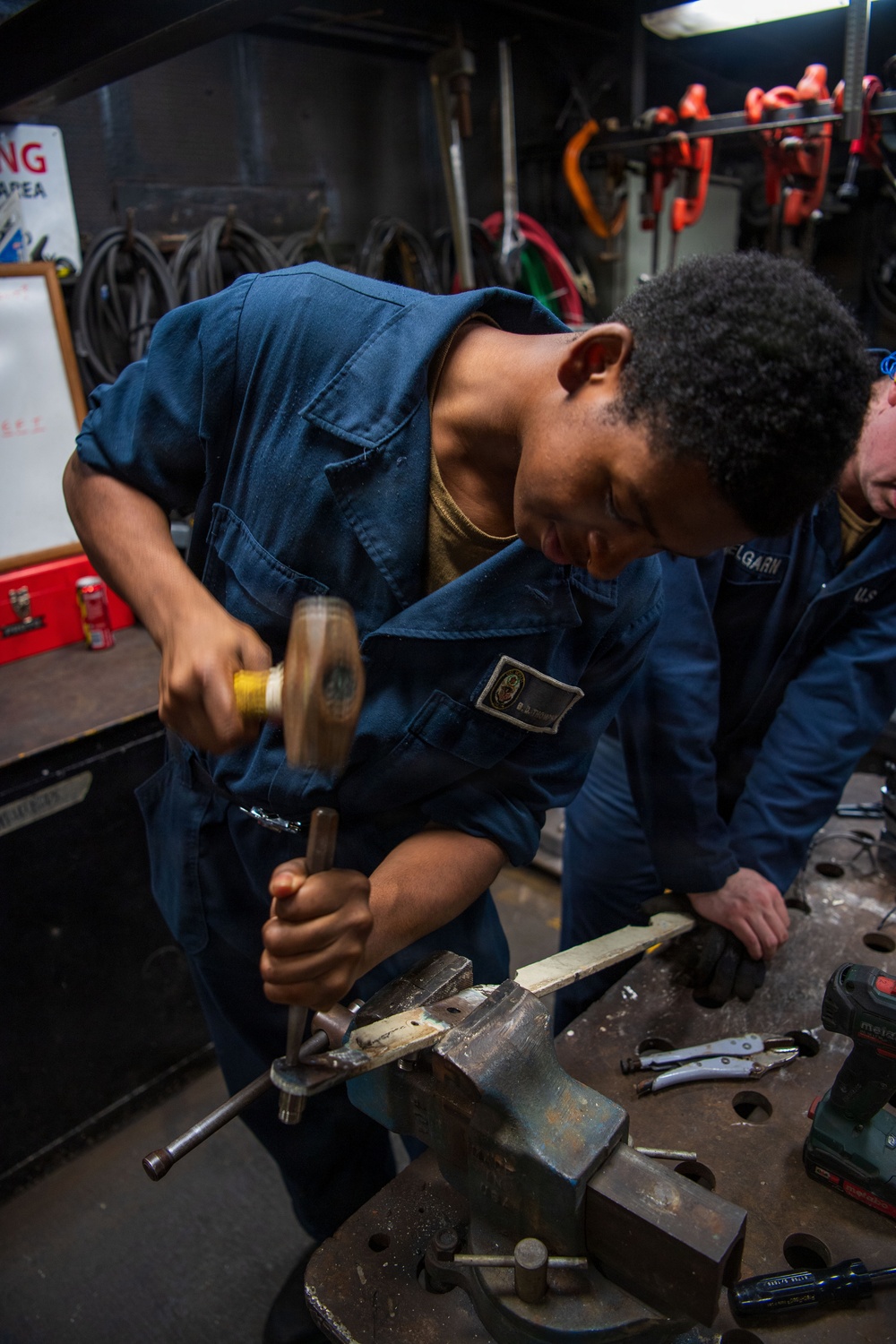 A young person wearing blue naval coveralls is using a hammer and chisel or punch on a metal piece secured in a bench vise. A second individual in similar blue U.S. Navy coveralls, with a partially visible name tag reading 'ELGARN', is assisting nearby. The setting appears to be a shipboard machine or repair shop with various tools and equipment visible in the background.